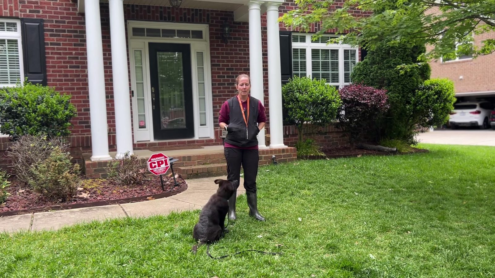 Handler standing at distance across a front lawn while a black German Shepherd puppy waits, during long-distance whistle recall training
